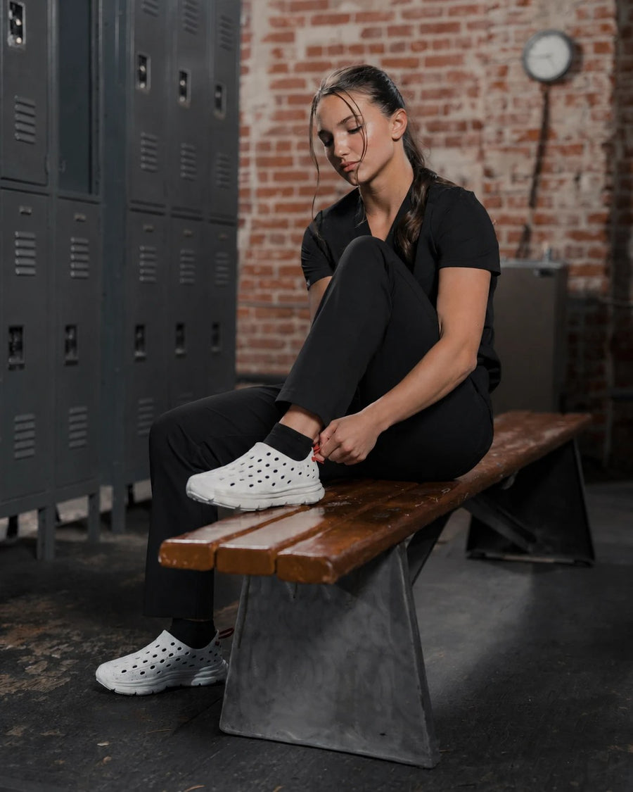 Healthcare worker in black scrubs on a locker room bench in Kane x Üni recovery shoes